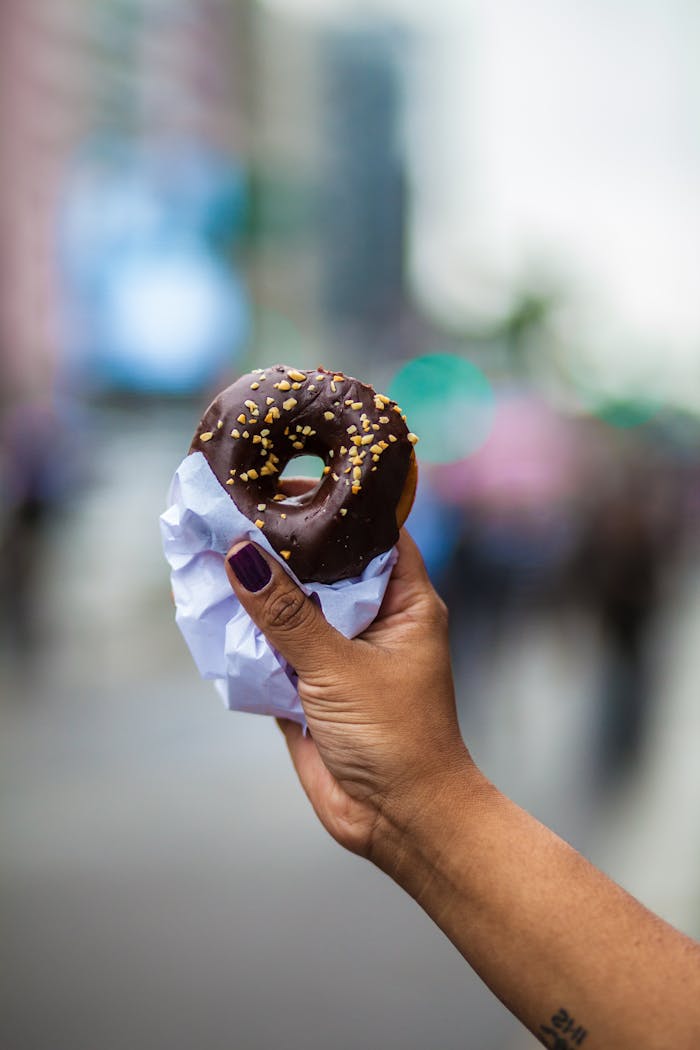 A person holds a chocolate glazed donut with sprinkles in an urban setting, showcasing food enjoyment.