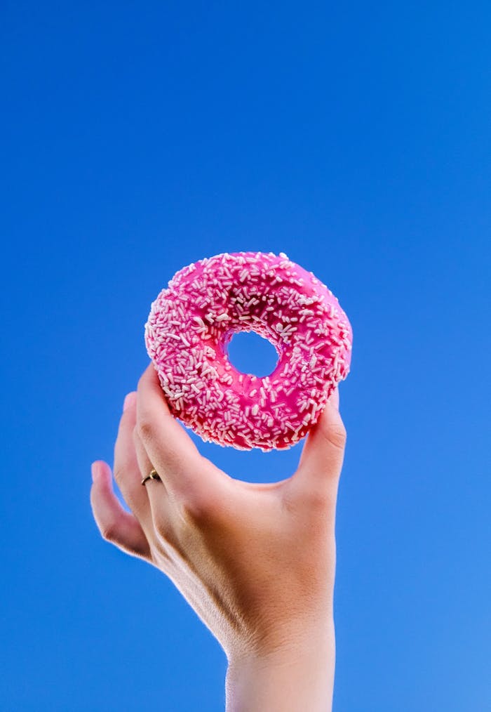 services-01 A hand holds a pink frosted donut with white sprinkles against a vibrant clear blue sky, offering a playful and colorful composition.