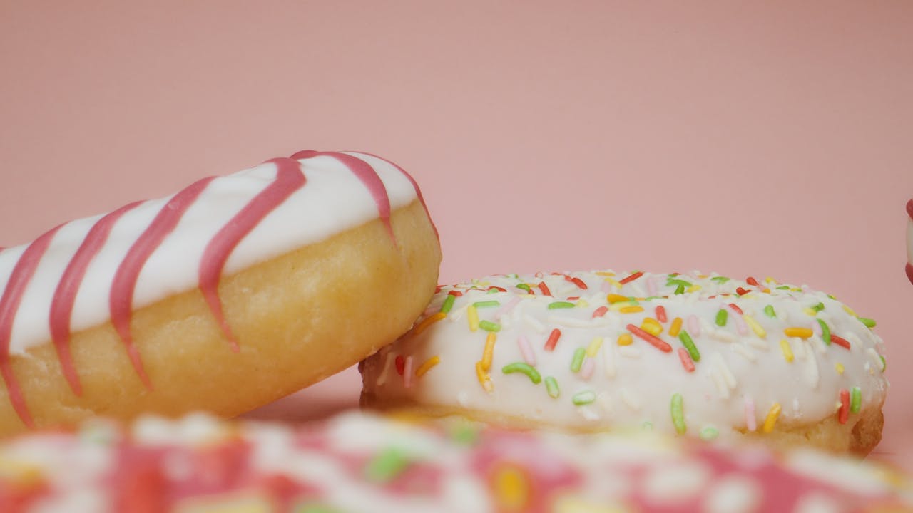 services-img Close-up of delicious donuts with colorful icing and sprinkles on a pink background.
