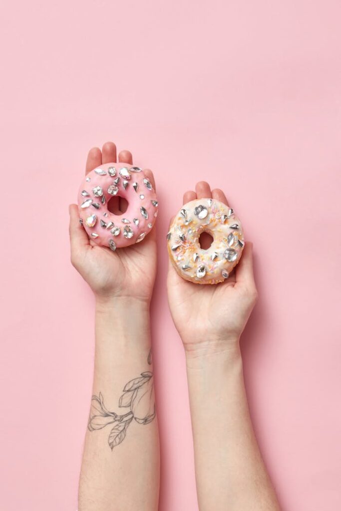 pexels photo 9228618 Close-up of tattooed hands holding pink and white donuts with colorful sprinkles.