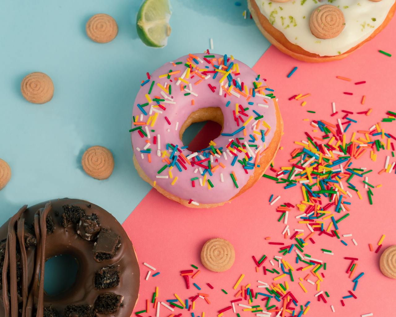 gallery-3 Top view of colorful donuts with sprinkles and biscuits on a two-tone background.